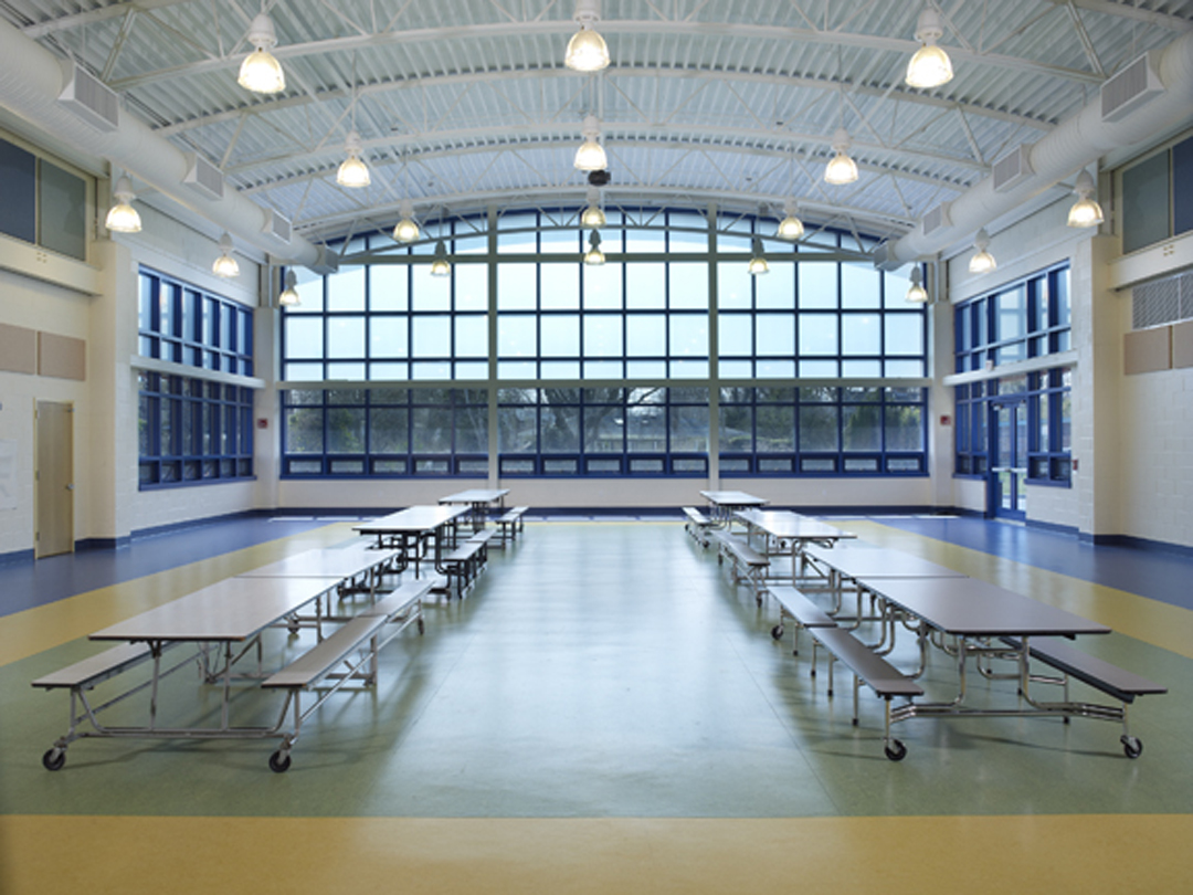 Empty school cafeteria with folded tables, large arched windows, and a high ceiling with hanging lights.