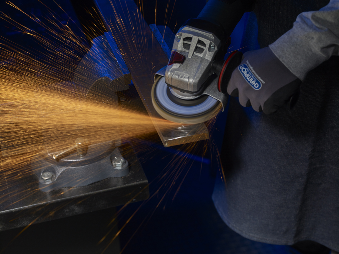 A person wearing safety gloves uses an angle grinder on a metal piece, emitting bright orange sparks in a dim workshop.