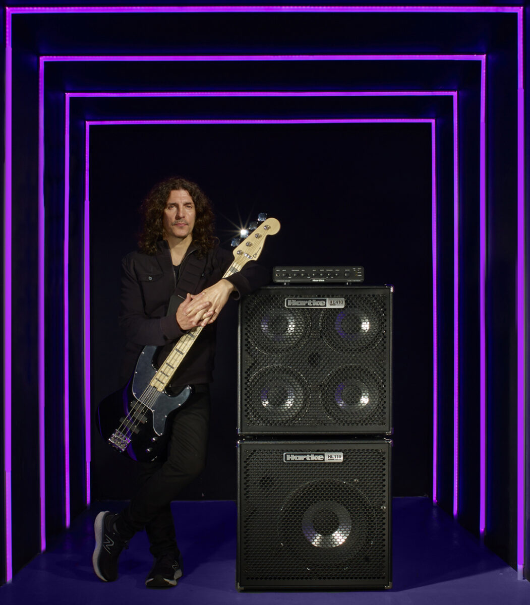 Man with curly hair standing and holding a bass guitar next to large speakers, framed by glowing neon lights.
